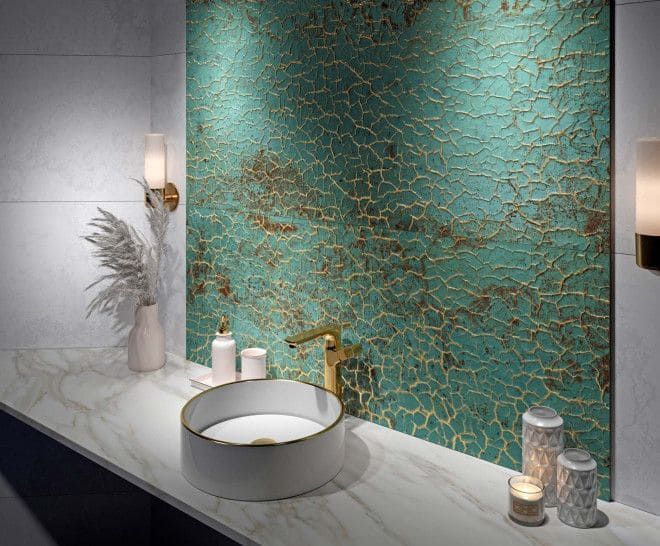 Baño con salpicadero de pintura agrietada en color verde azulado y dorado, lavabo blanco y grifo dorado sobre una encimera de mármol.