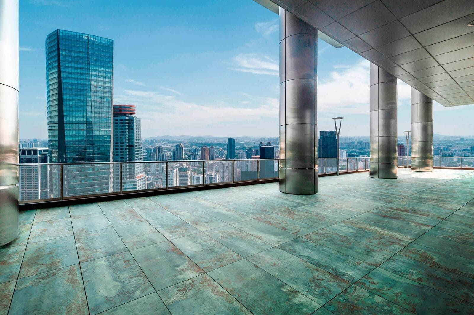 El horizonte de la ciudad visto desde una terraza al aire libre con grandes columnas de metal y suelos de baldosas de color azul verdoso bajo un cielo azul.