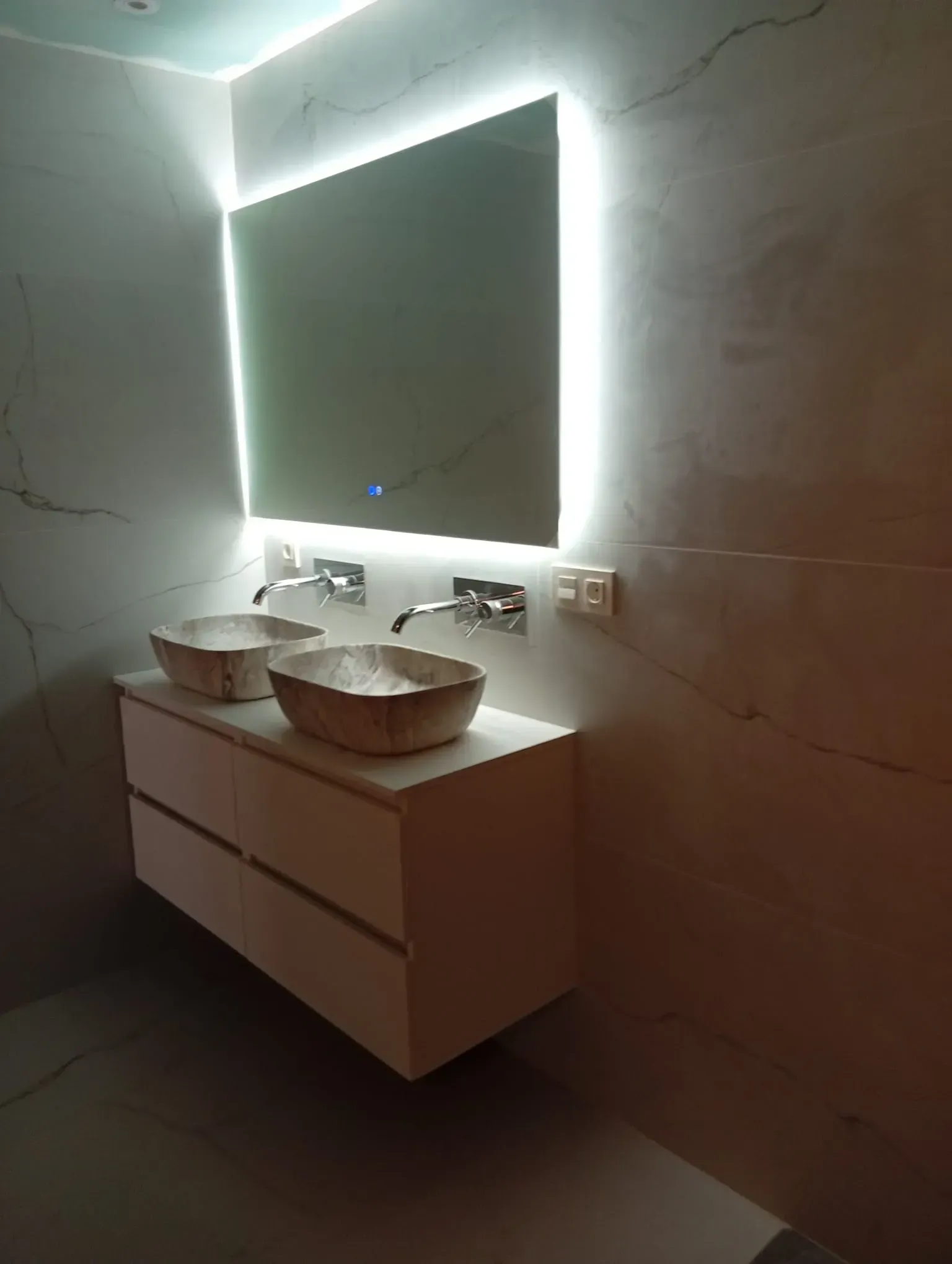 Tocador de baño moderno con dos lavabos sobre encimera, espejo iluminado y paredes estilo mármol.