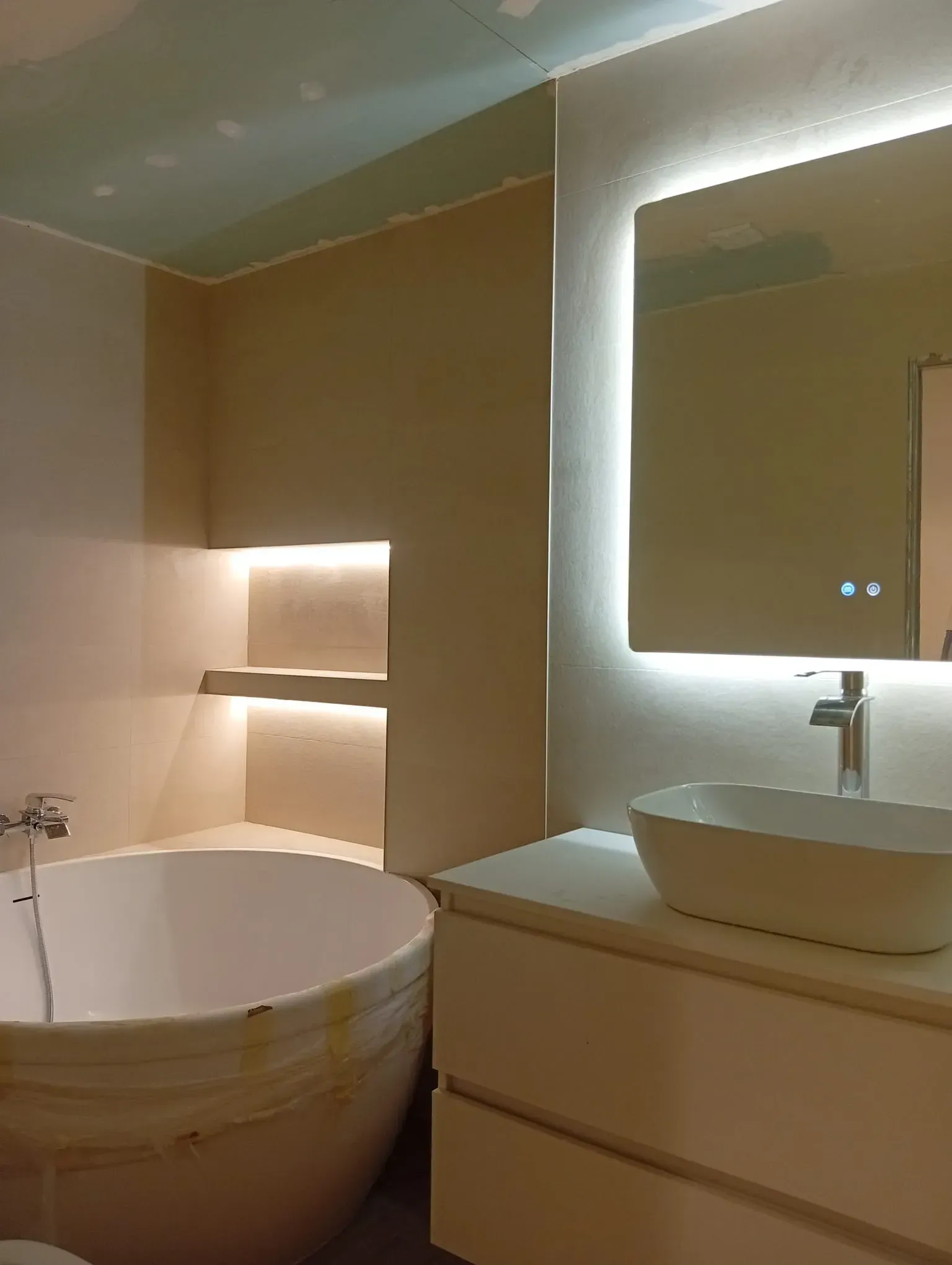 Baño moderno con bañera independiente, lavabo con tocador y espejo iluminado en tonos beige cálidos.