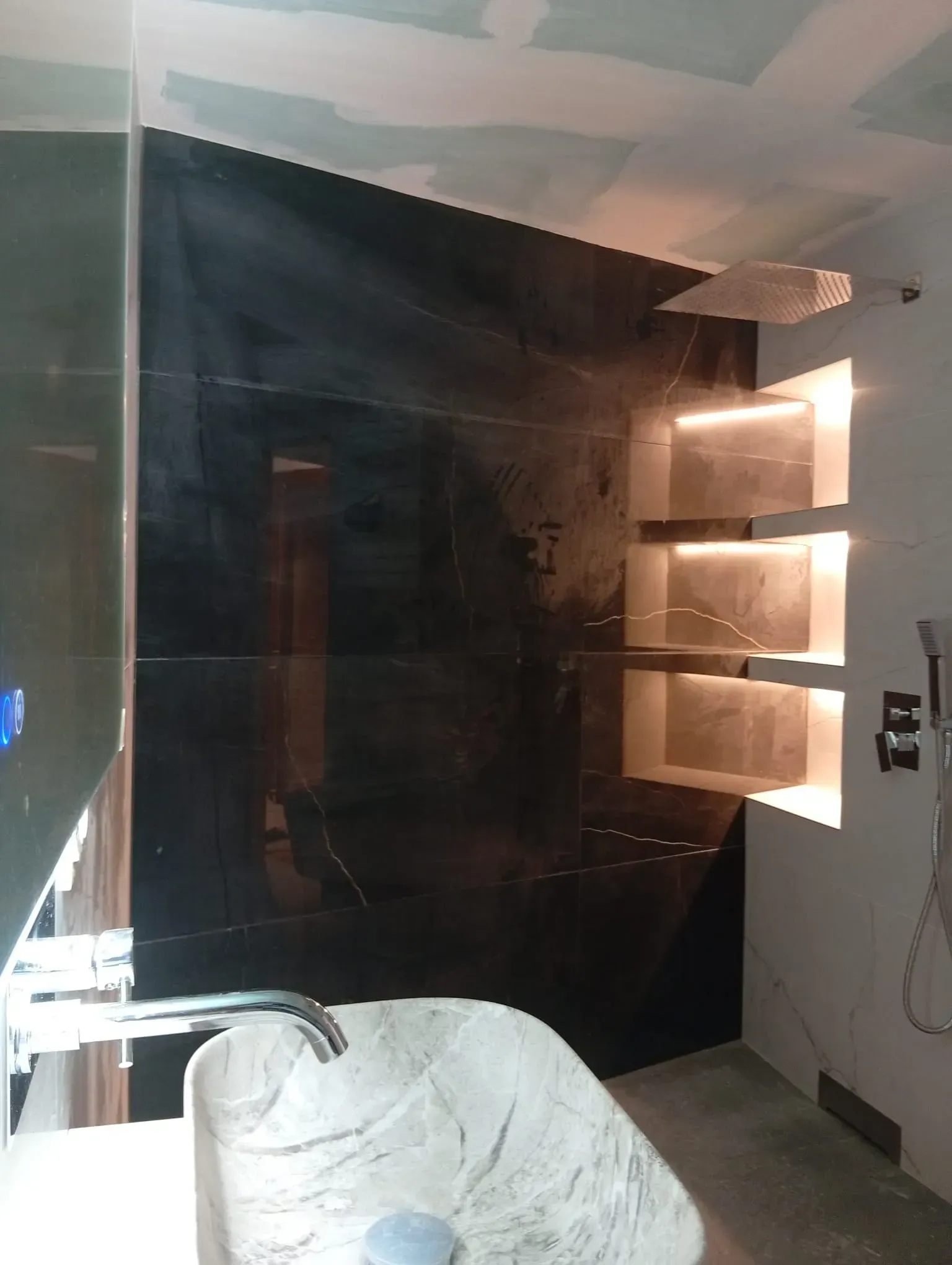 Baño con poca luz, pared negra, lavabo con tocador, espejo y estantes iluminados a la derecha.