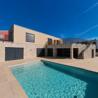 Casa moderna de color beige con detalles de piedra y una piscina turquesa bajo un cielo azul claro.