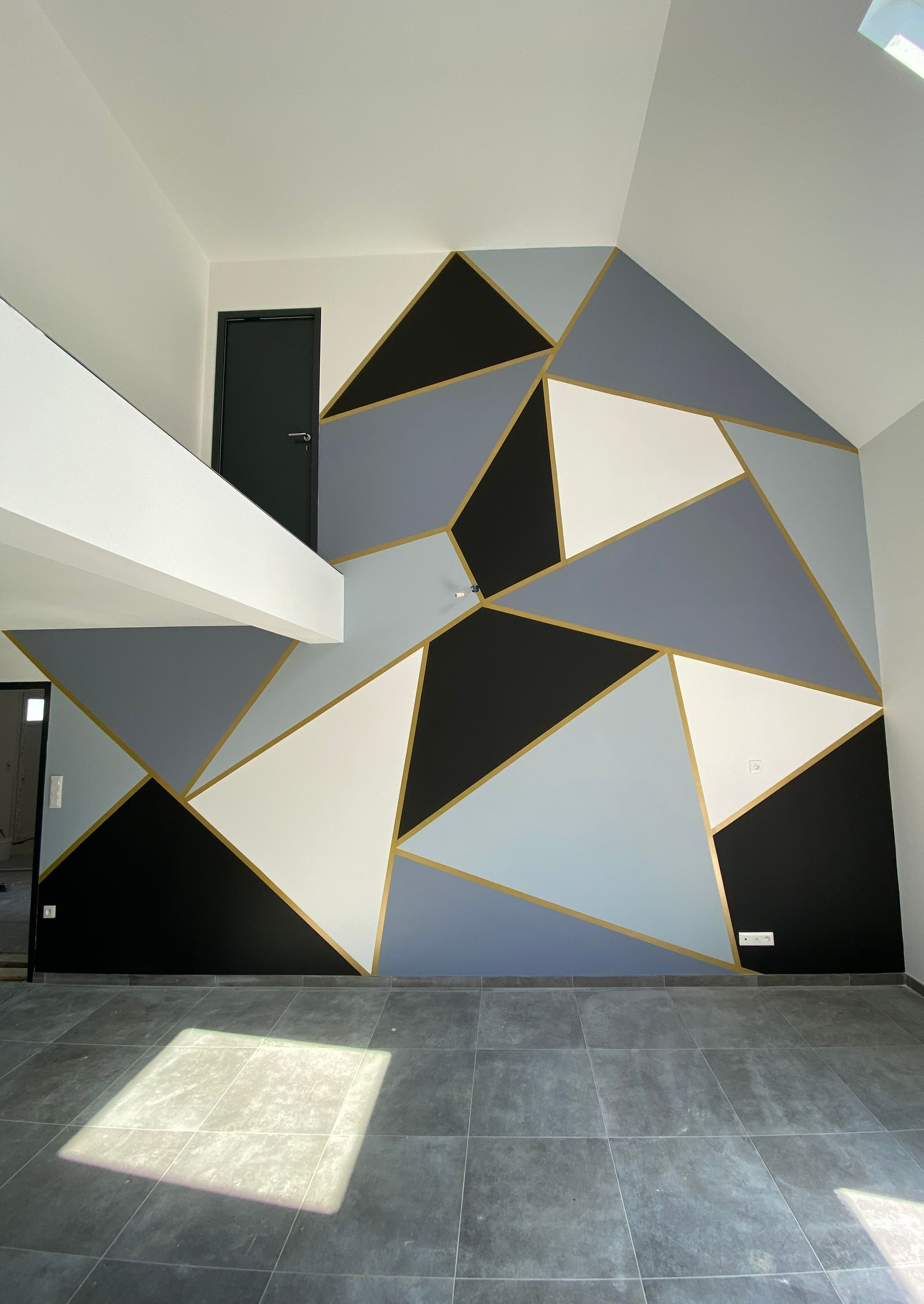 Décoration murale géométrique : triangles gris, noirs et blancs avec bordure dorée dans une pièce au sol foncé et à la porte foncée.