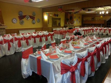 Salón de banquetes preparado para un evento formal con sillas blancas, lazos rojos y mantelería.