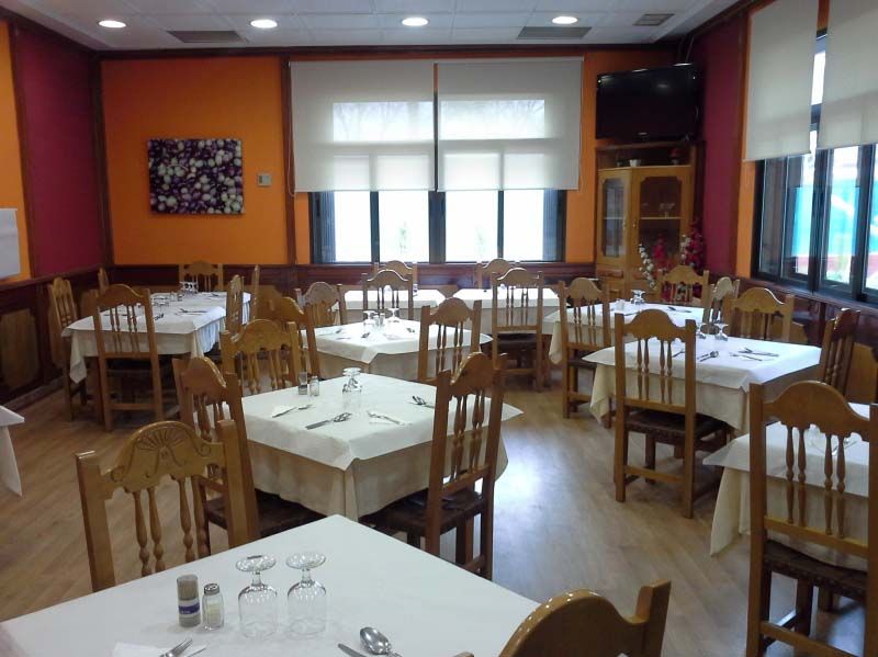Comedor vacío de un restaurante con sillas de madera, mesas con manteles blancos y paredes rojas con grandes ventanales.