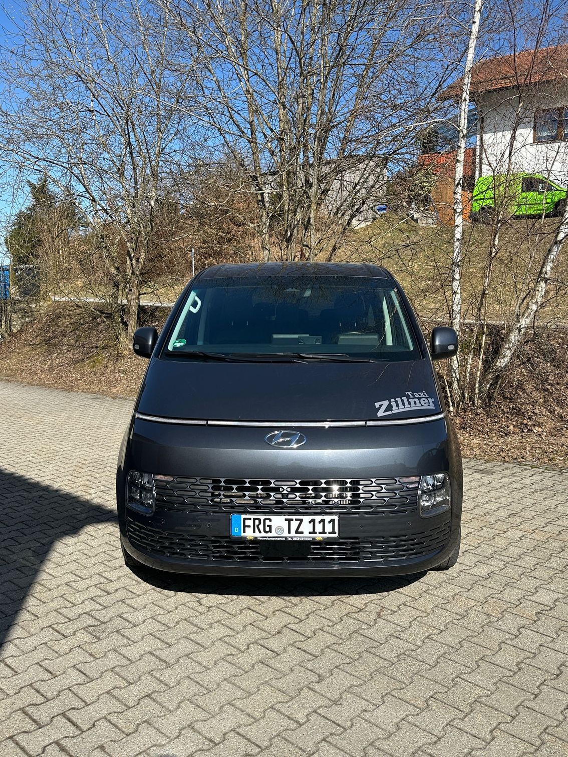 Nahaufnahme der Front eines dunklen Fahrzeugs (Taxi), umgeben von dichtem Geäst kahler Bäume bei sonnigem Wetter.