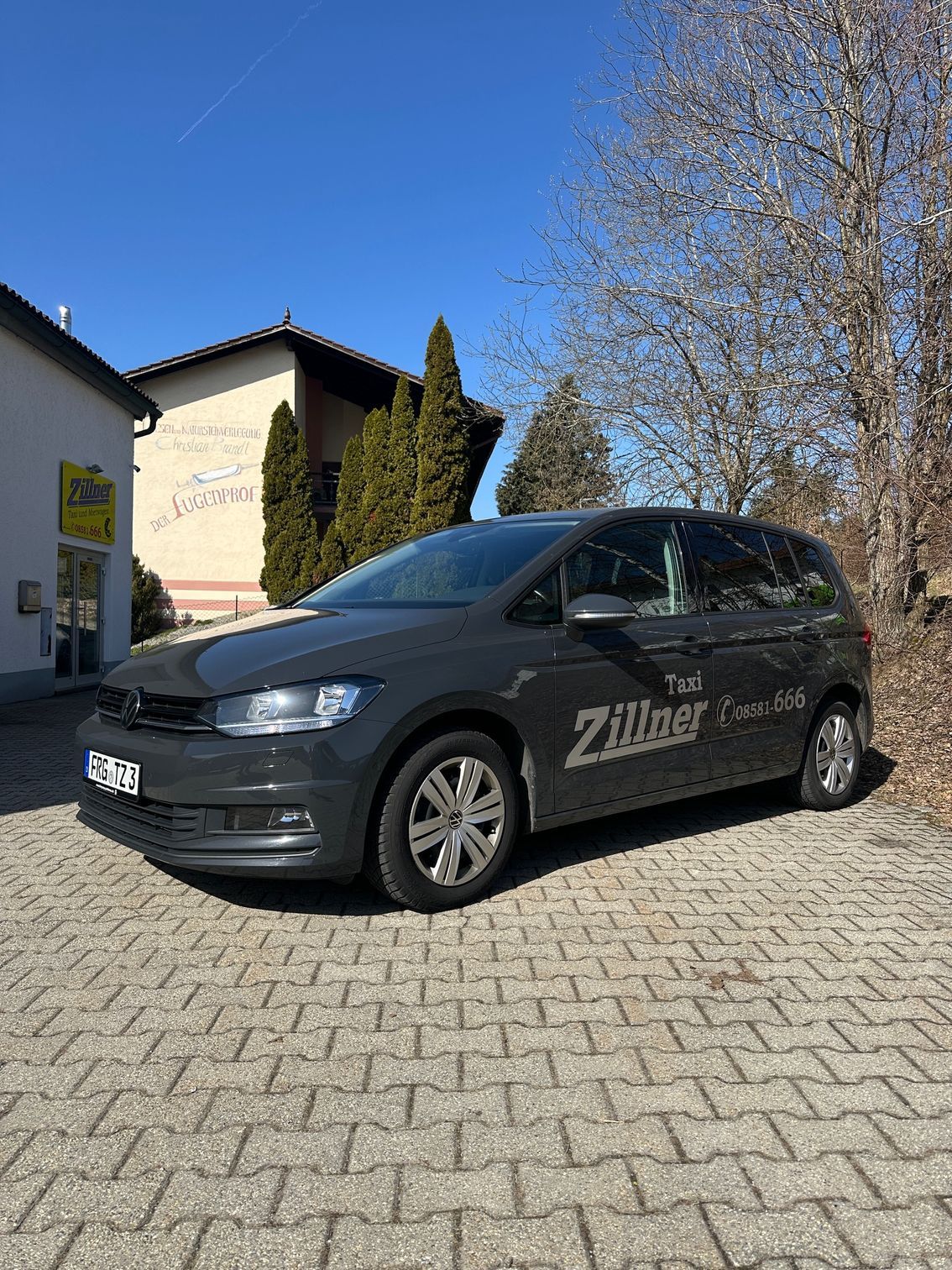 Seitenansicht eines dunklen Taxis vor einem weißen Firmengebäude mit gelbem Logo unter strahlend blauem Himmel.