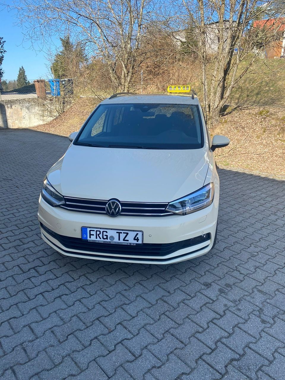 Frontalansicht eines hellen Taxis mit gelbem Taxischild auf dem Dach, geparkt auf einem Platz vor  Bäumen.