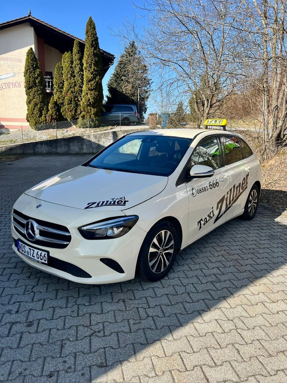 Helles Taxi von der Seite vor Firmengebäude und hohen Zypressen.