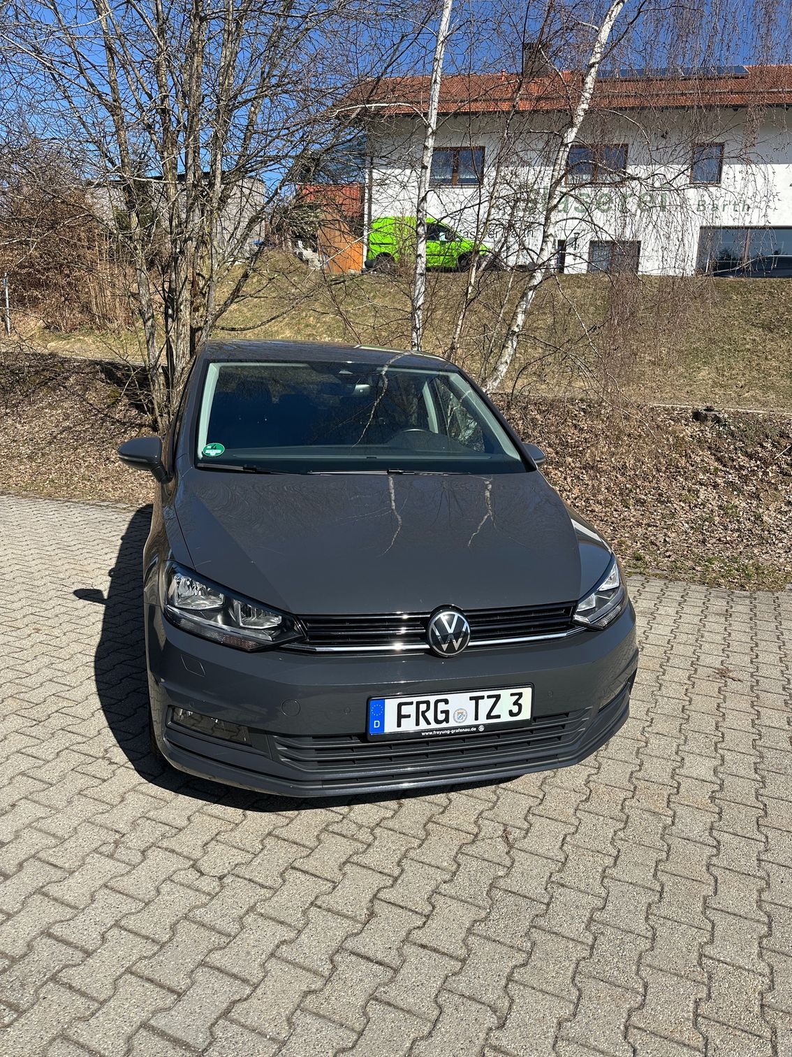 Frontale Nahaufnahme eines dunklen Taxis, das vor einer Böschung mit kahlen Bäumen und einem Wohnhaus im Hintergrund parkt.