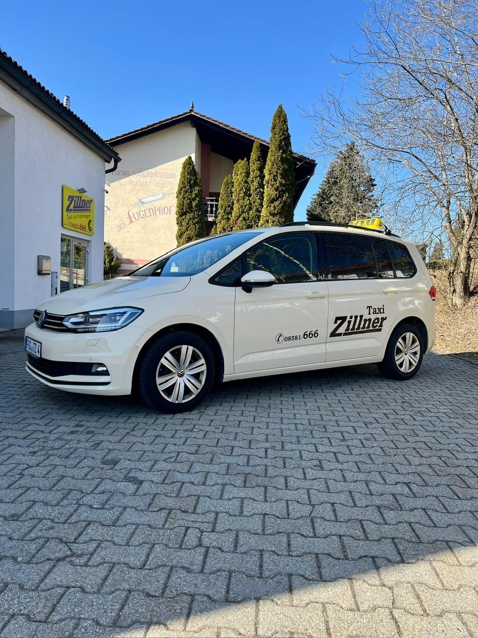 Seitenansicht eines hellen Taxis vor dem Firmengebäude mit der Aufschrift „Taxi“. Im Hintergrund sind grüne Zypressen.