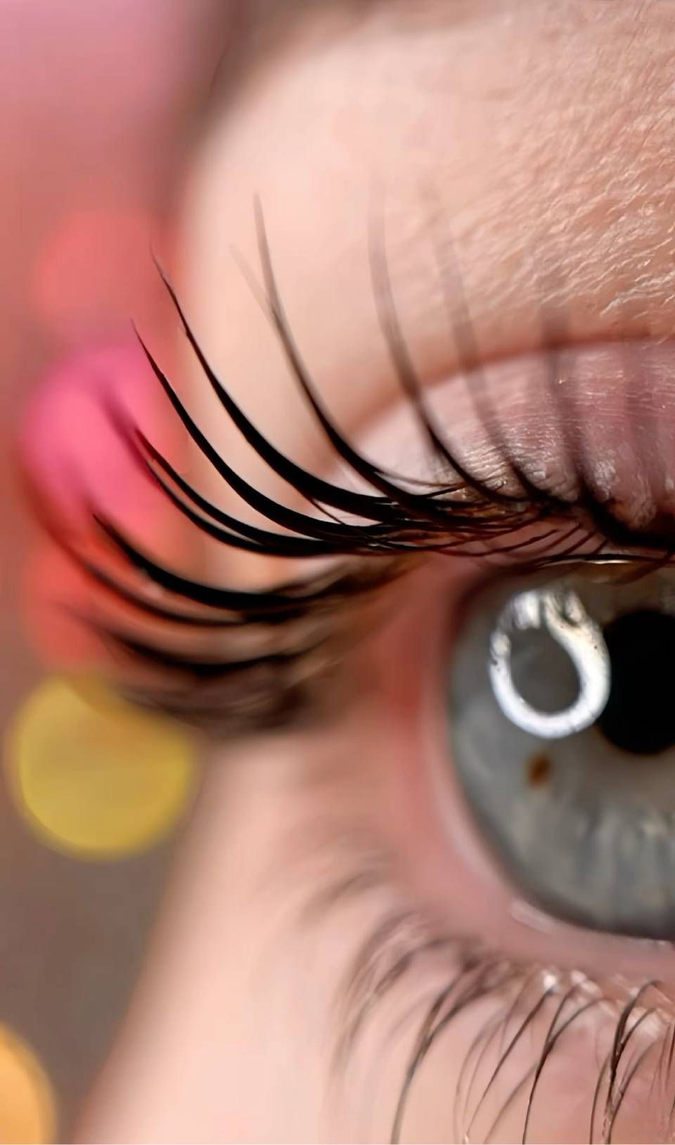 Nahaufnahme eines menschlichen Auges mit langen, schwarzen Wimpern und blauer Iris, im Hintergrund ein verschwommener, farbenfroher Bokeh-Effekt.