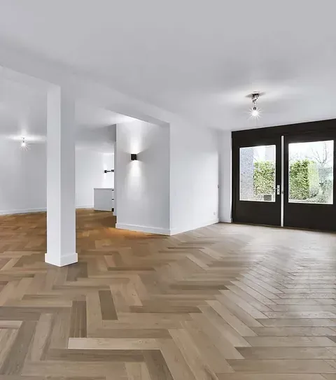 Habitación vacía con piso de madera en espiga, paredes blancas y puertas dobles negras que dan a un jardín.