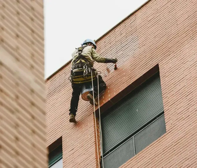 Persona con equipo de seguridad limpiando pared de ladrillos, suspendida por cuerdas, al lado de una ventana.
