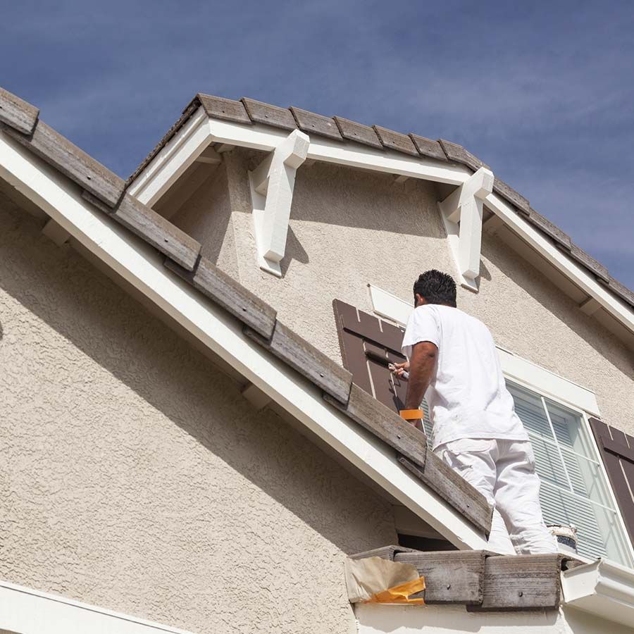 Hombre pintando el exterior de una casa con contraventanas marrones, estuco beige y molduras blancas.