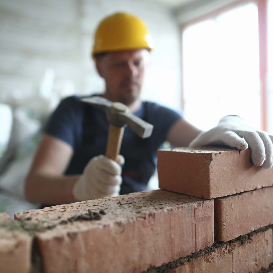 Un albañil que lleva un casco amarillo coloca ladrillos con un martillo y una mano enguantada de color blanco.