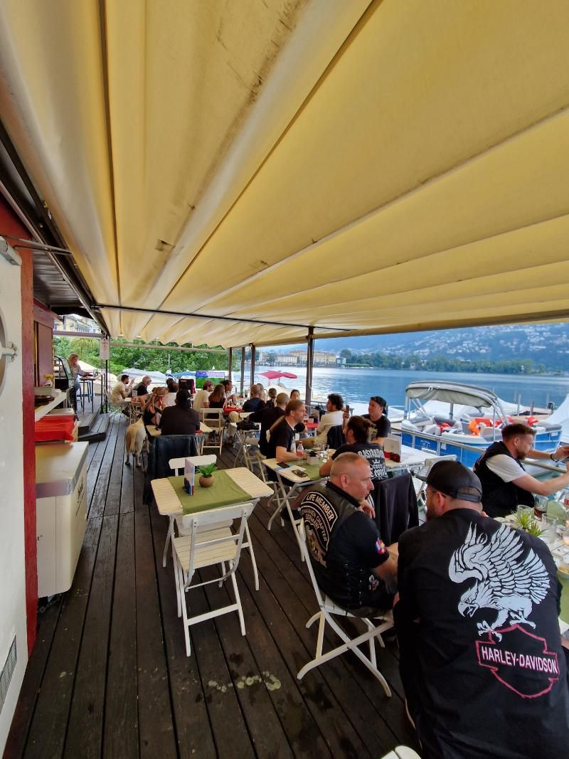 Un gruppo di persone è seduto a dei tavoli sotto una tettoia che si affaccia su un lago.