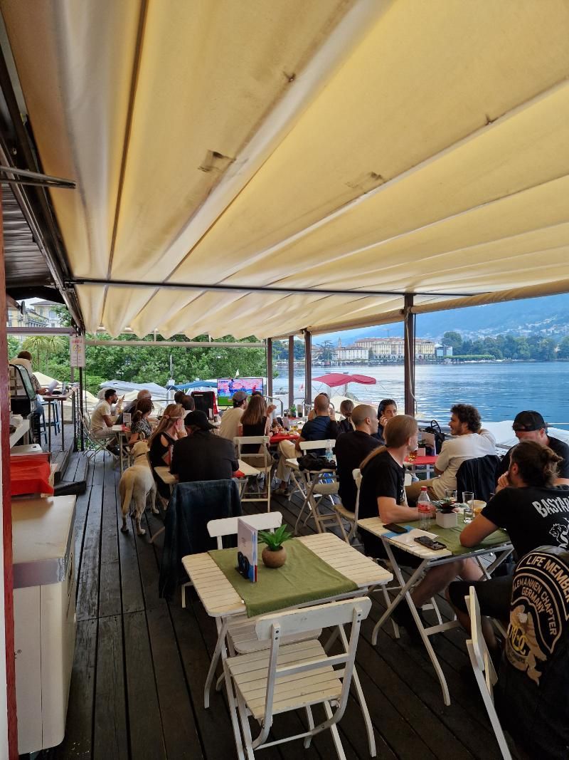 Un gruppo di persone è seduto a dei tavoli sotto una tettoia che si affaccia su uno specchio d'acqua.
