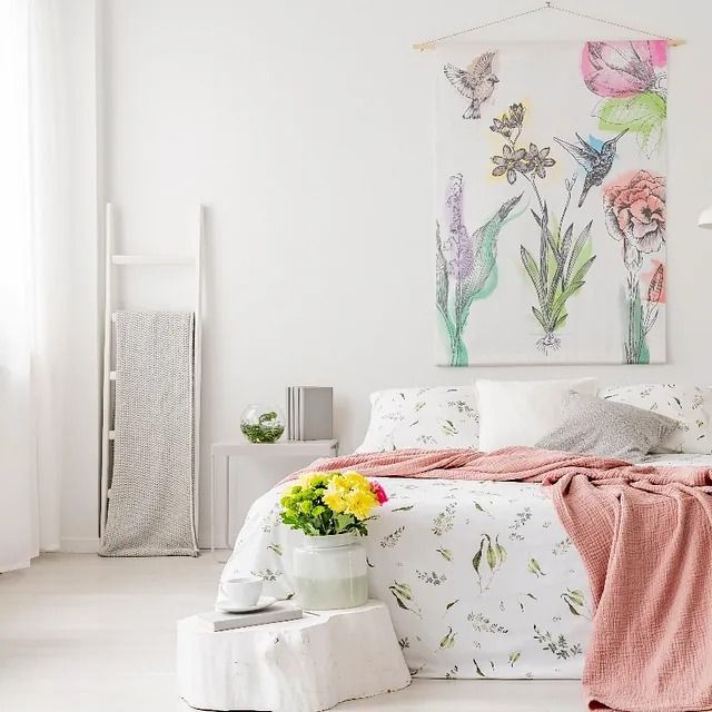 Habitación blanca luminosa con ropa de cama floral, manta rosa y cuadro en la pared sobre la cama.