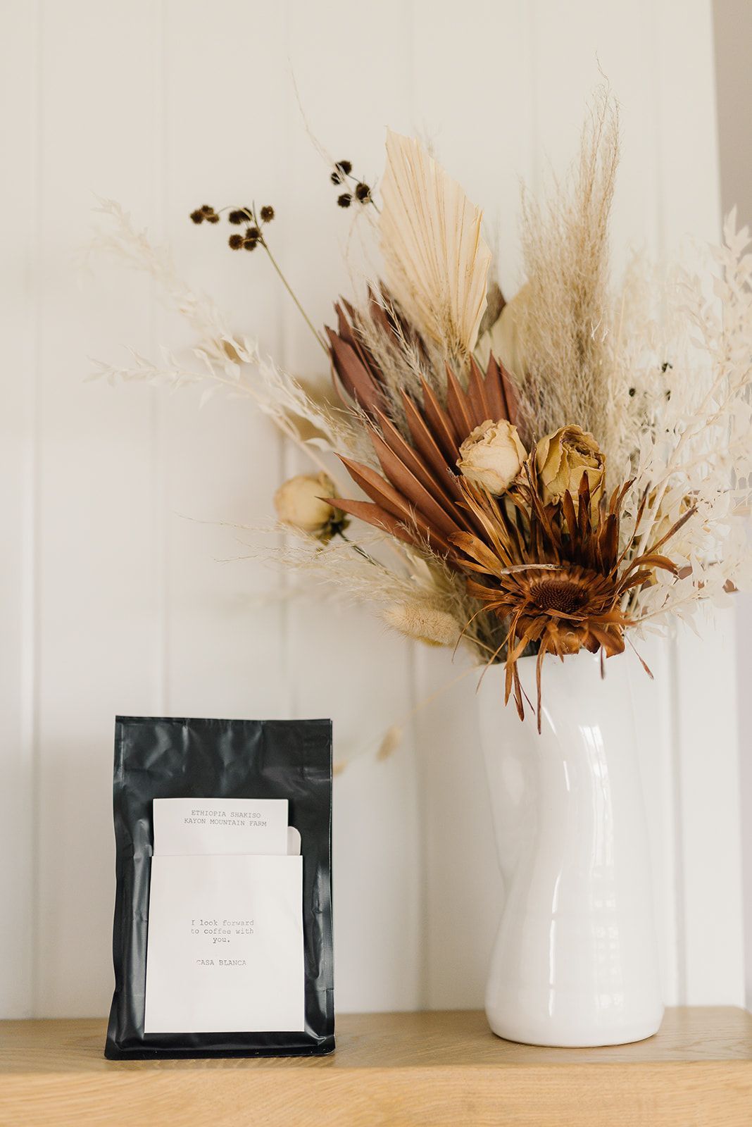 A bag of coffee and a vase of dried flowers are on a shelf.