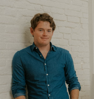 Person in a blue button-up shirt standing against a white brick wall, smiling softly.
