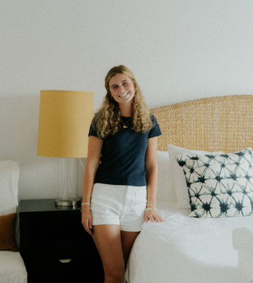 Woman standing beside a bed in a bedroom, wearing a dark shirt and white shorts.