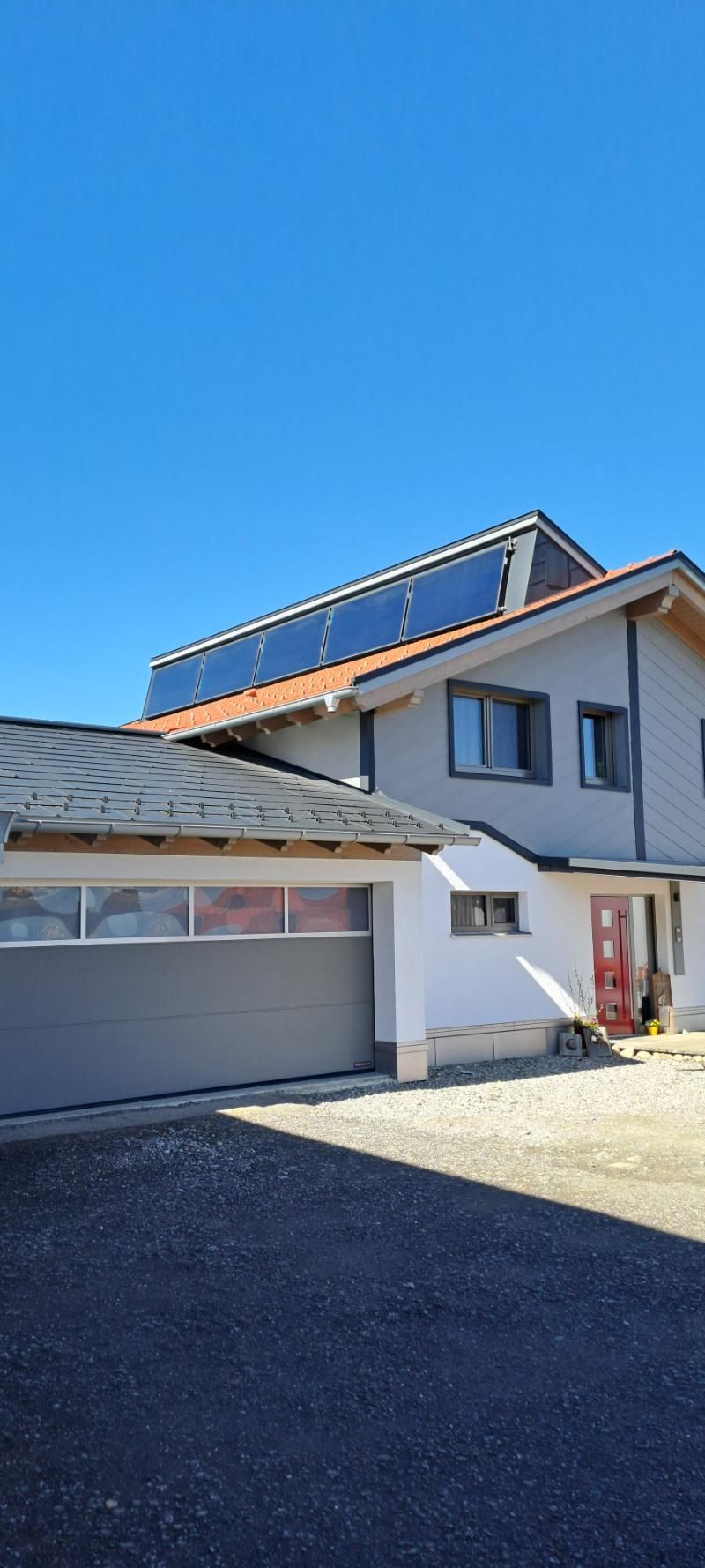 Ein modernes zweistöckiges Haus mit Garage, roten Dachziegeln und Solarpaneelen vor einem klaren blauen Himmel.