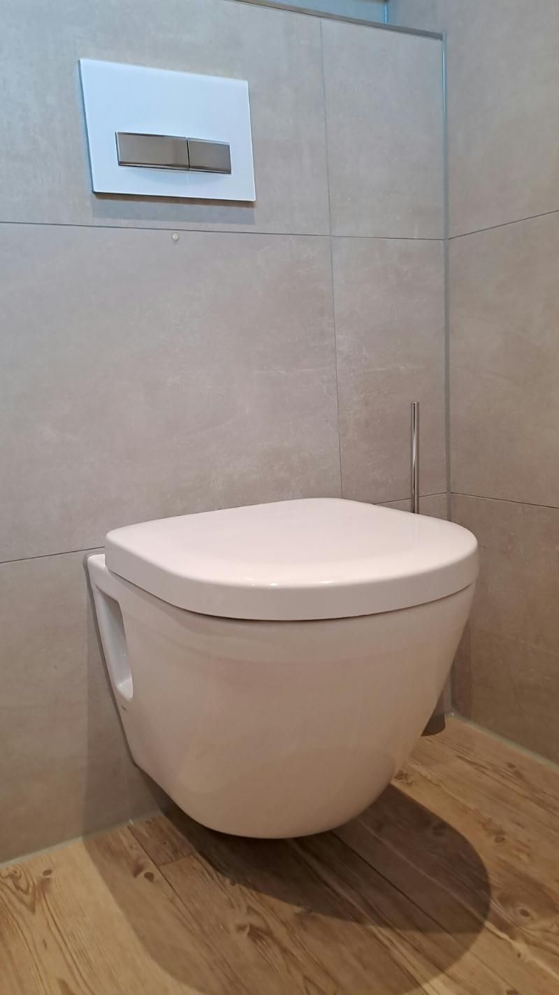 Eine moderne, weiße, wandhängende Toilette mit geschlossenem Deckel vor beigefarbenen Fliesenwänden und Holzboden.