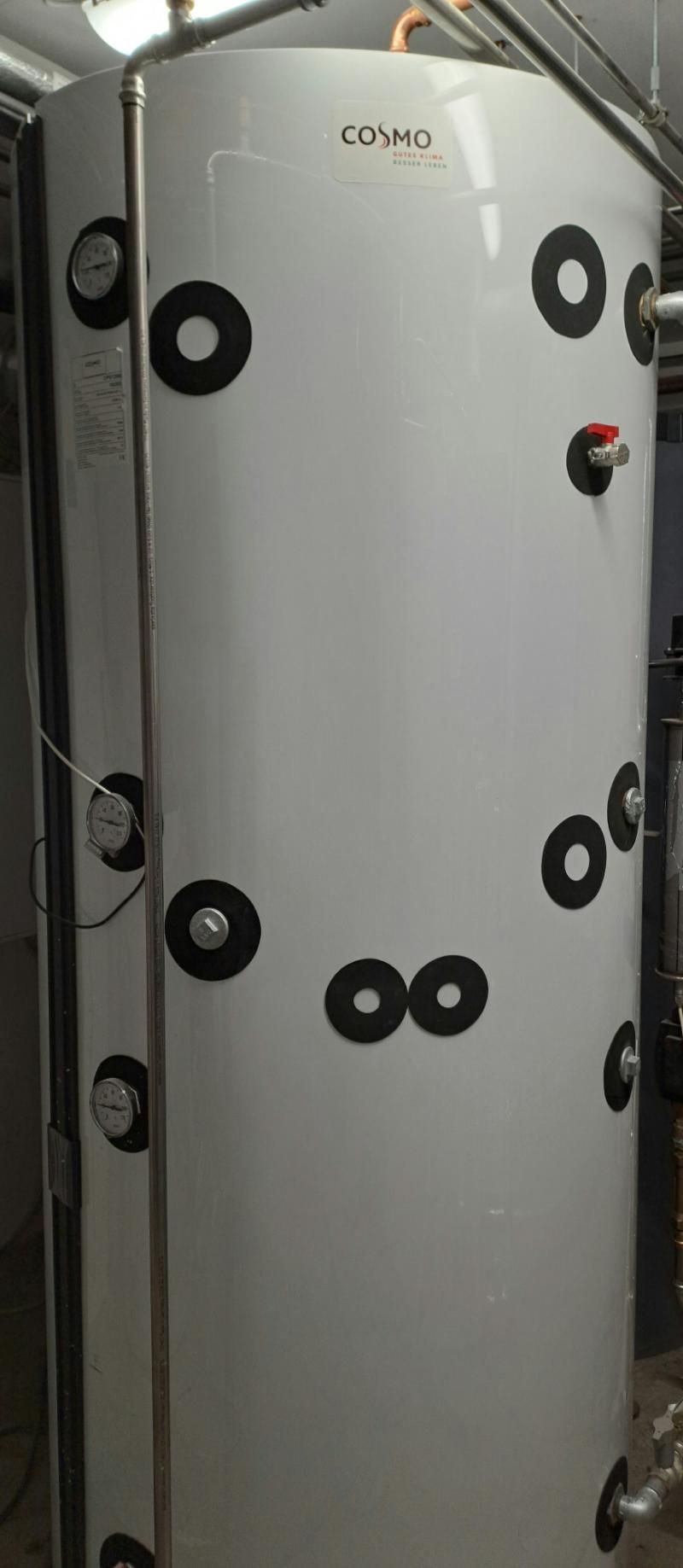 Weißer industrieller Warmwasserspeicher mit mehreren schwarzen Befestigungspunkten und gut sichtbaren Manometern in einem Hauswirtschaftsraum.