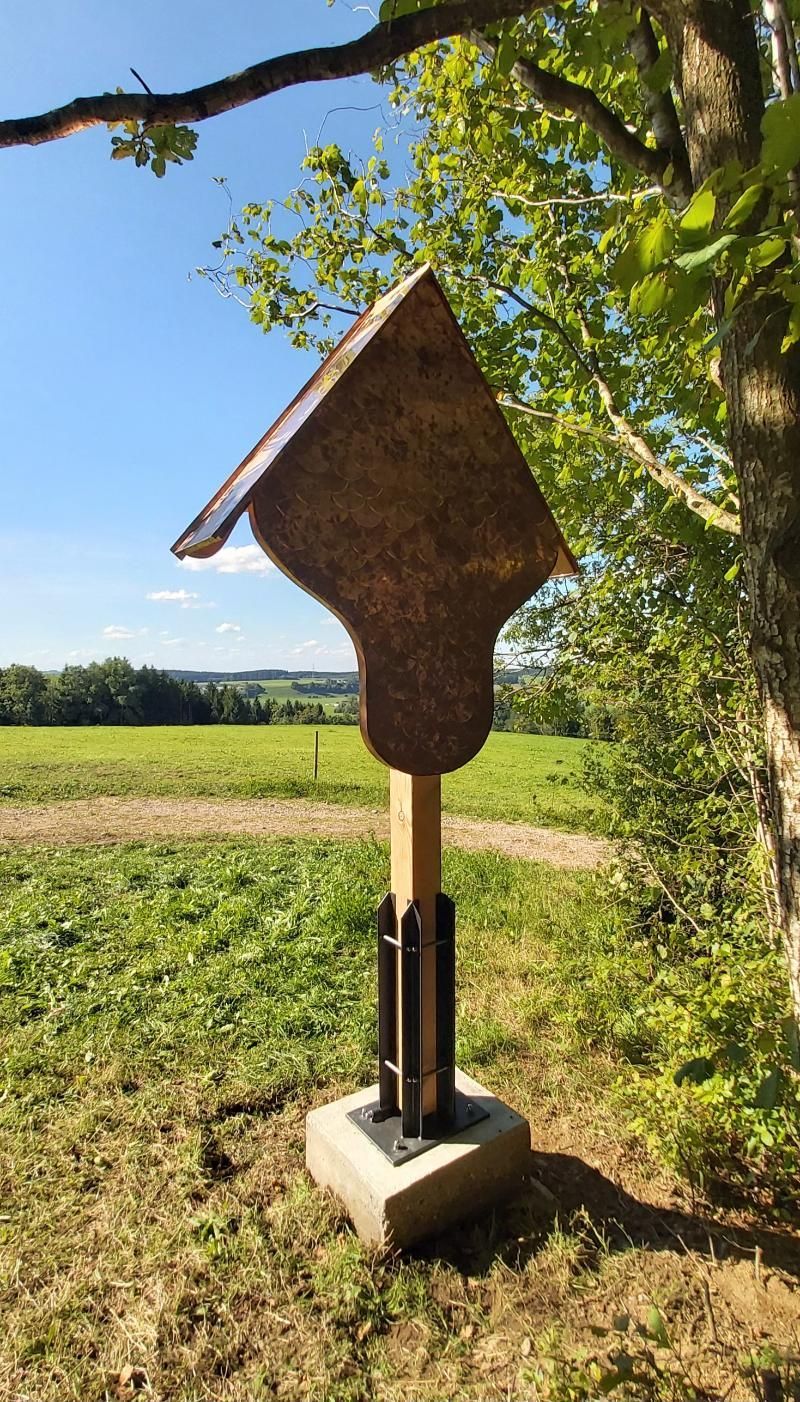 Auf einem Steinsockel in einer grasbewachsenen Wiese unter klarem, blauem Himmel steht ein moderner Wegschrein aus Metall.