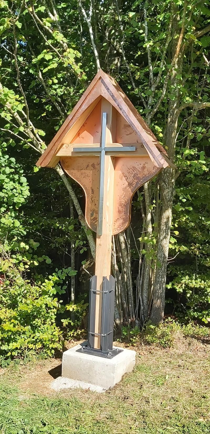 Ein modernes Wegkreuz mit Holzrahmen und Metallkreuz, aufgestellt auf einem Steinsockel in einem üppigen, grünen Waldgebiet.