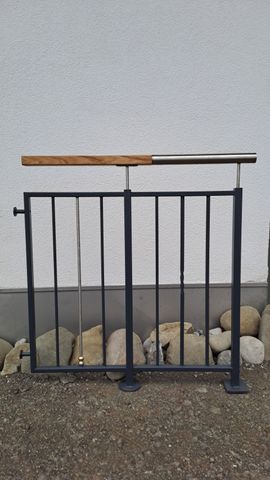 Ein Werkzeug mit Holzgriff aus Metall liegt auf einem schwarzen Metalltor vor einer Steinmauer, unter der sich Felsen befinden.