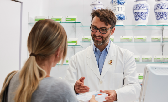 Farmacista in camice bianco che assiste un cliente al bancone della farmacia; sullo sfondo scaffali di medicinali.