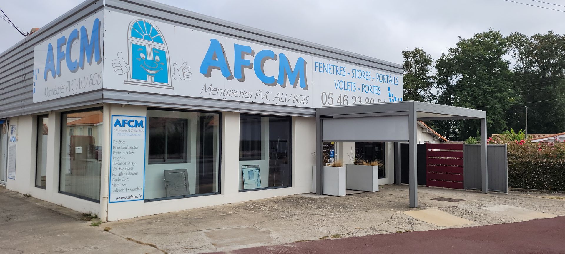 Façade du magasin AFCM avec enseigne bleue et blanche ; toit gris, fenêtres et zone pavée devant.