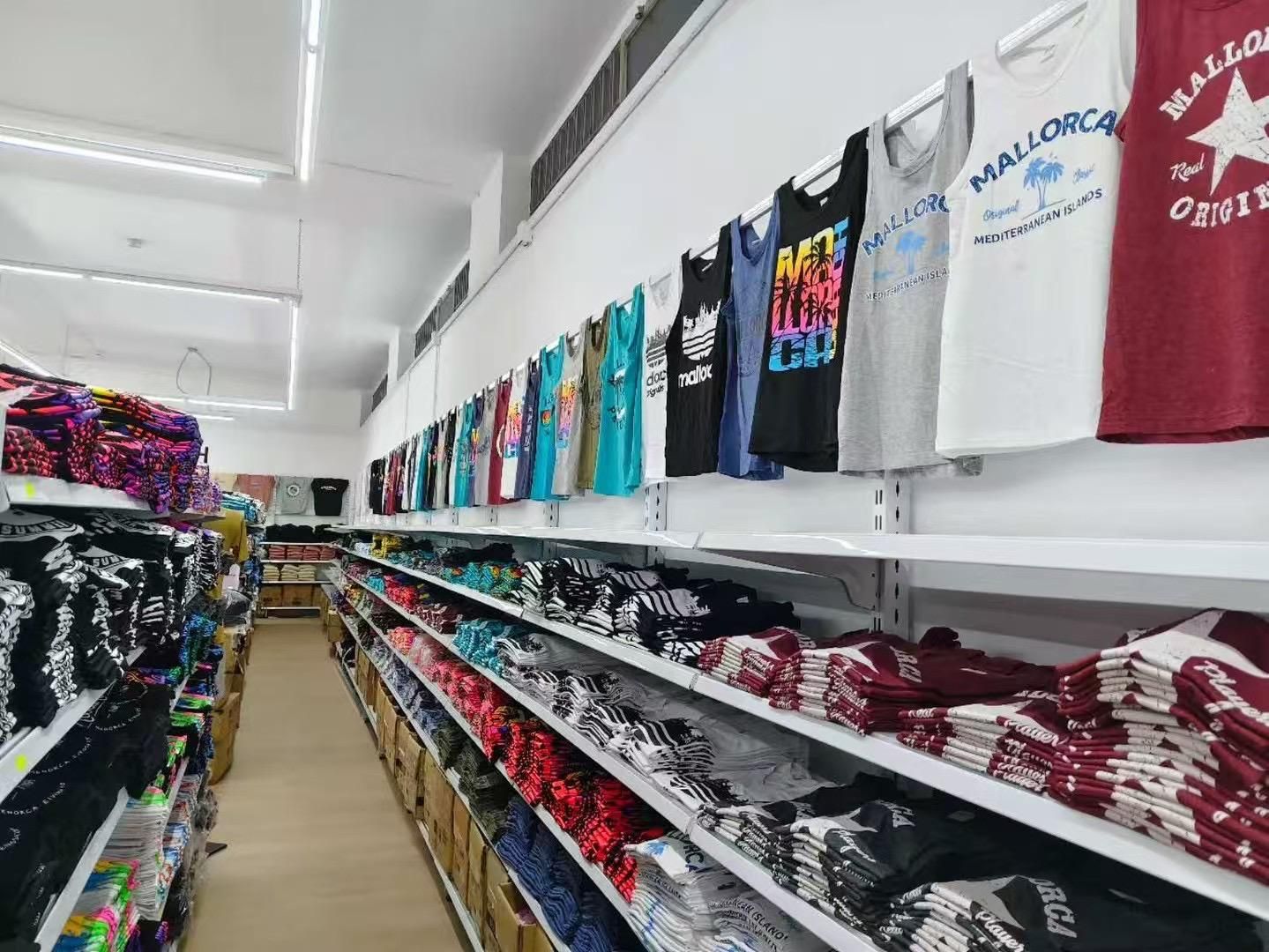 Filas de ropa en estanterías y colgadas en una pared de una tienda, incluidas camisetas con estampados de Mallorca.