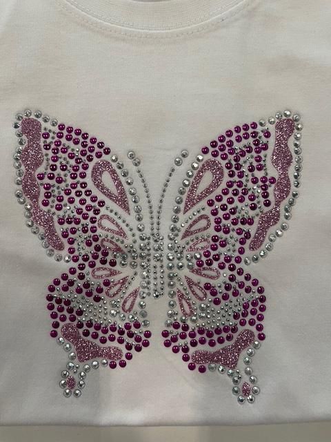 Camiseta blanca con diseño de mariposa de pedrería en tonos rosa y plata.