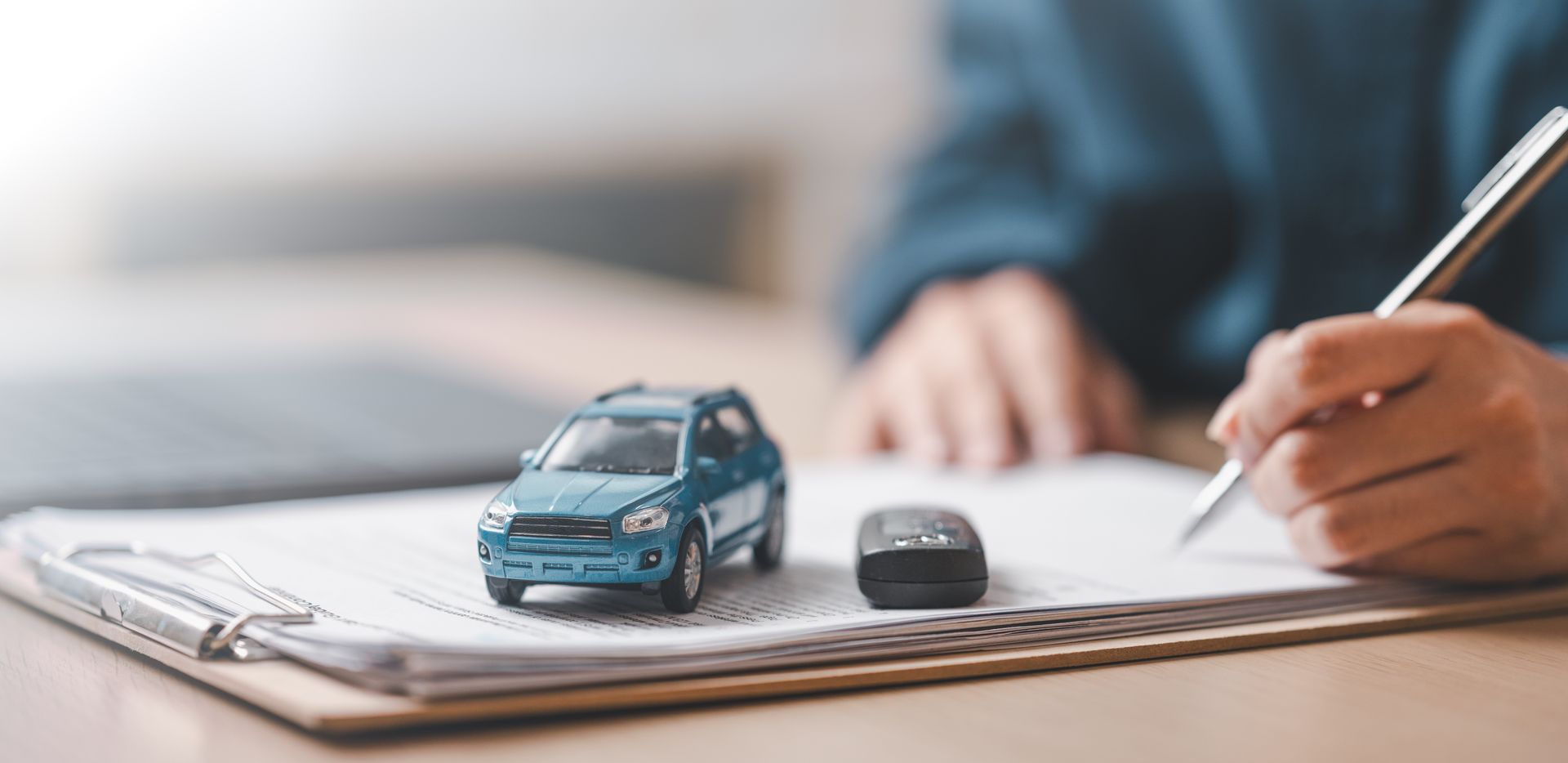 Une voiture miniature et une clé sont posées sur un porte-documents pendant qu'une personne écrit au stylo à un bureau