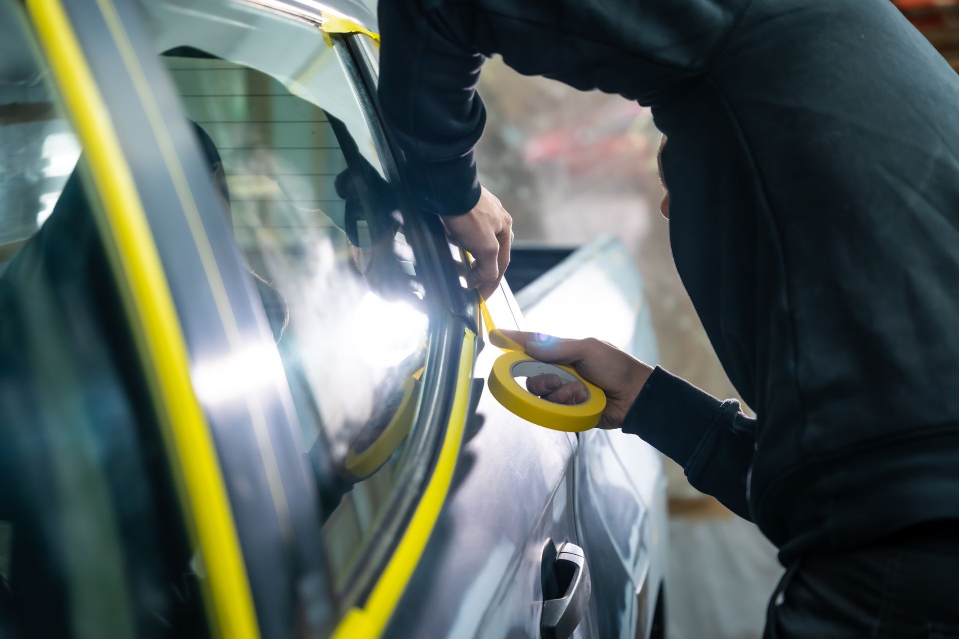 Des mains appliquent du ruban adhésif jaune sur une portière de voiture dans un atelier avec des reflets brillants sur le véhicule.