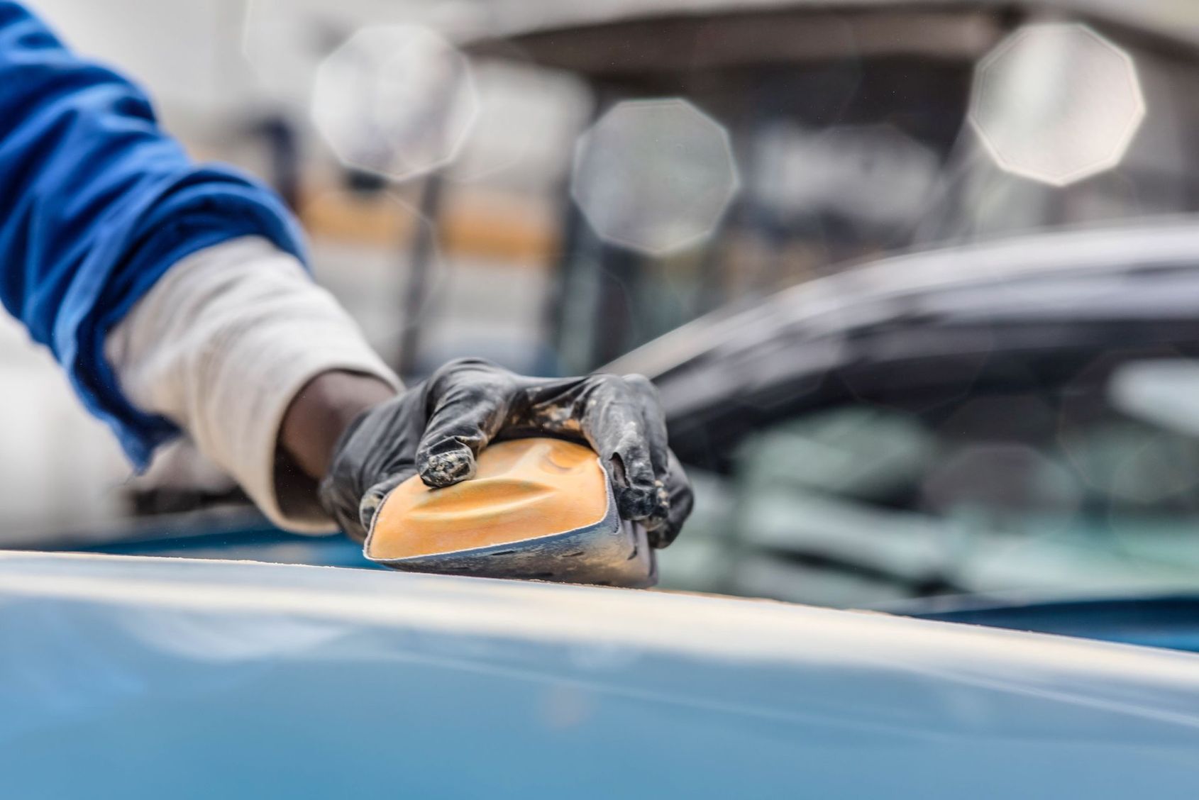 Main gantée polissant le capot bleu d'une voiture avec une éponge dans un atelier automobile