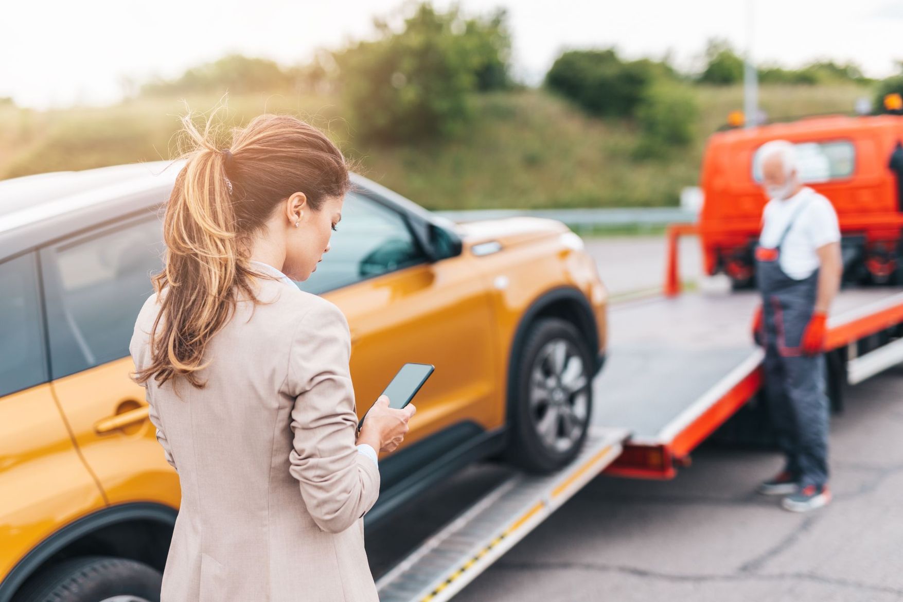 Un dépanneur charge un SUV orange sur une dépanneuse au bord d'une route pendant qu'une femme consulte son téléphone à proximité.