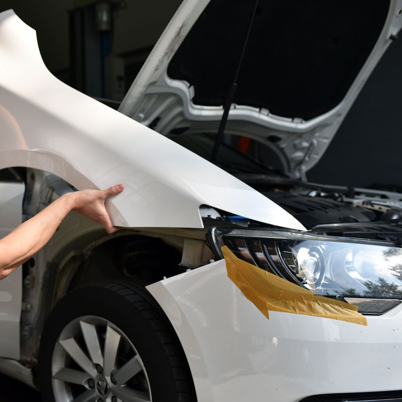 Une personne colle du ruban adhésif sur l'aile avant endommagée d'une voiture blanche près du capot ouvert dans un atelier de réparation.