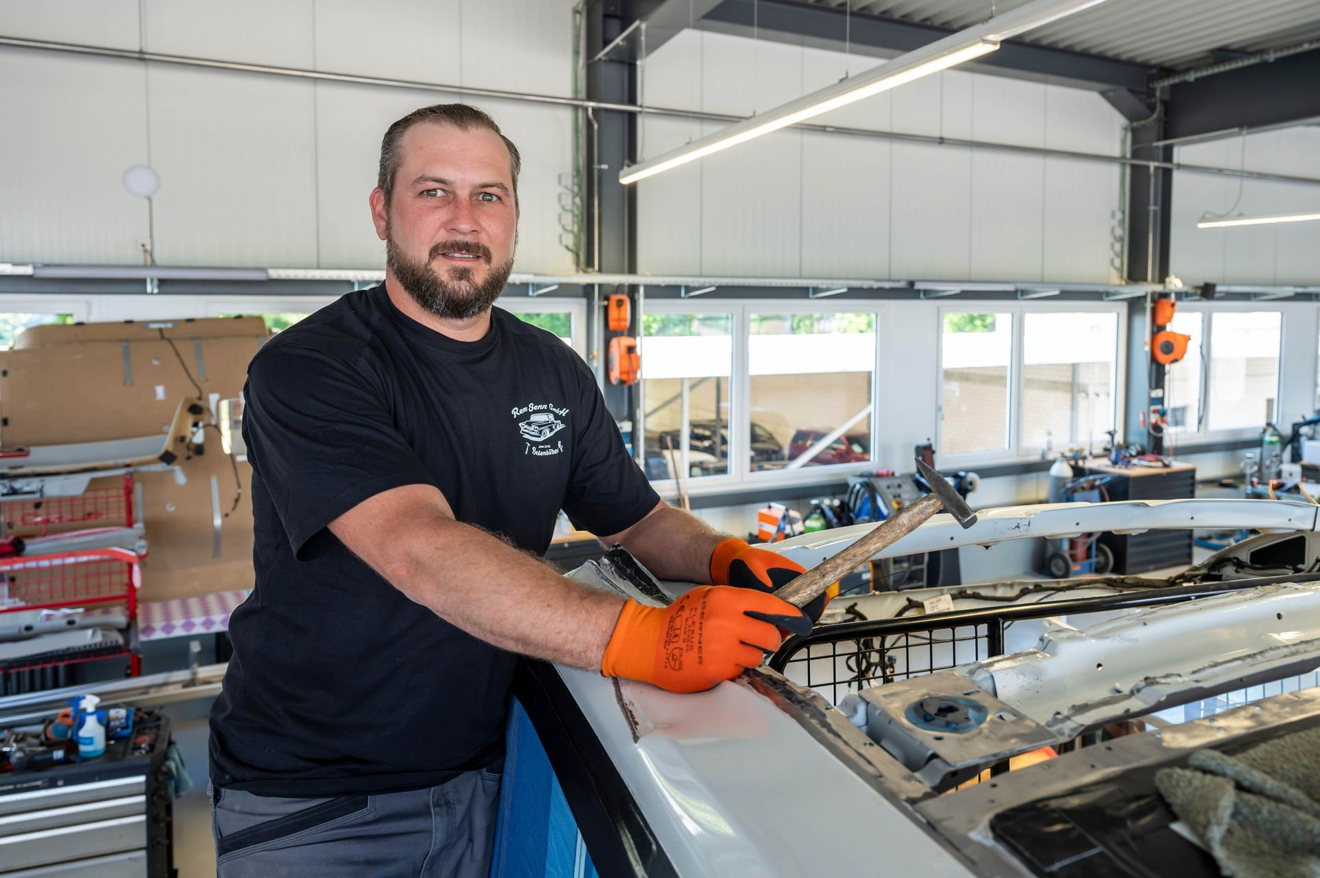 Mann in schwarzem Hemd, orangefarbenen Handschuhen, mit Hammer in der Hand, arbeitet in einer Reparaturwerkstatt an einem Auto.