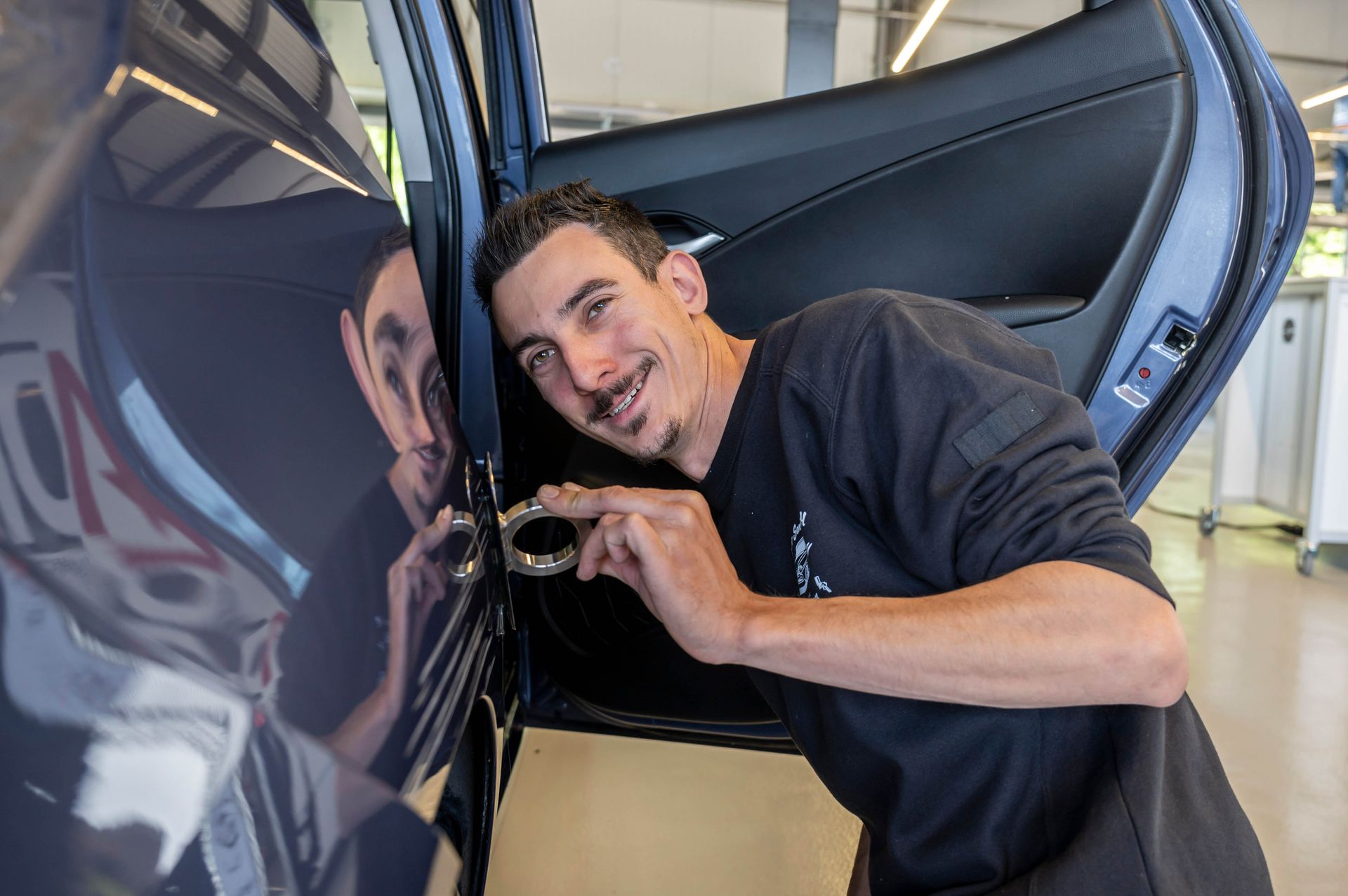 Mechaniker arbeitet lächelnd an einer Autotür. Dunkelhaariger Mann in schwarzem Hemd beugt sich in einer Werkstatt über ein dunkles Auto.