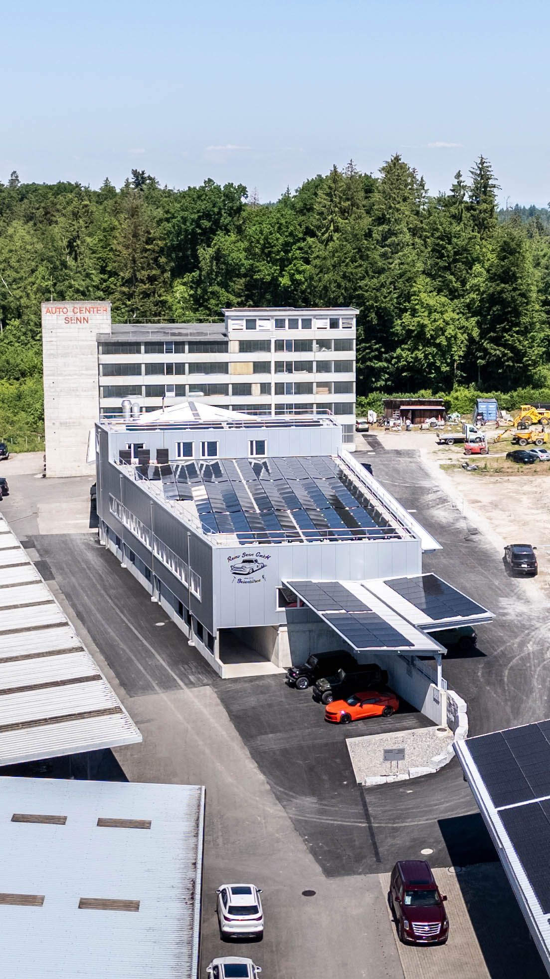 Modernes Gebäude mit Solarzellen auf dem Dach, draußen geparkte Autos, umgeben von Bäumen.