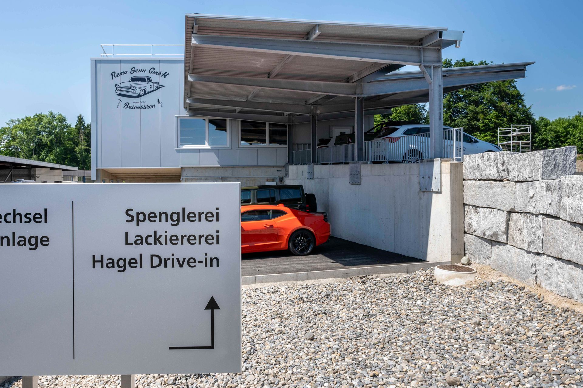 Ein orangefarbener Sportwagen fährt in eine Autowerkstatt. Schild weist auf „Hagel Drive-in“ hin.
