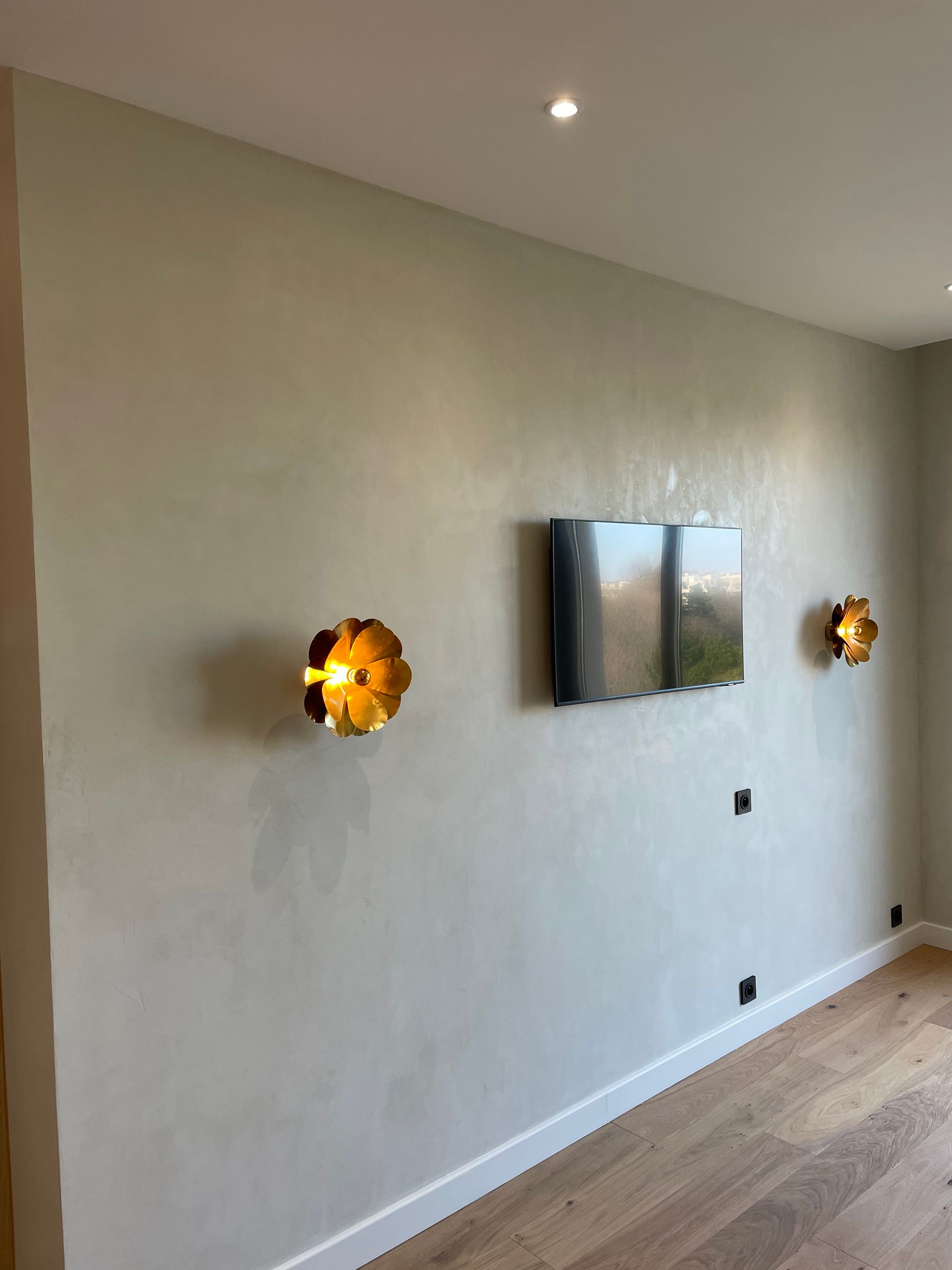 Mur clair orné de deux appliques murales dorées en forme de fleur et d'un téléviseur fixé au mur. Sol en bois.
