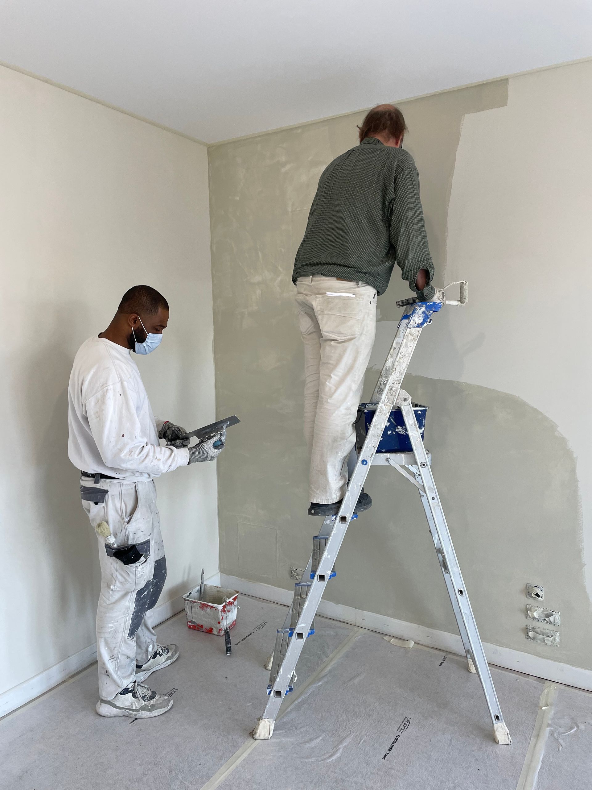Deux peintres enduisent un mur dans une pièce. L'un est sur une échelle, l'autre debout ; tous deux portent des vêtements de travail.