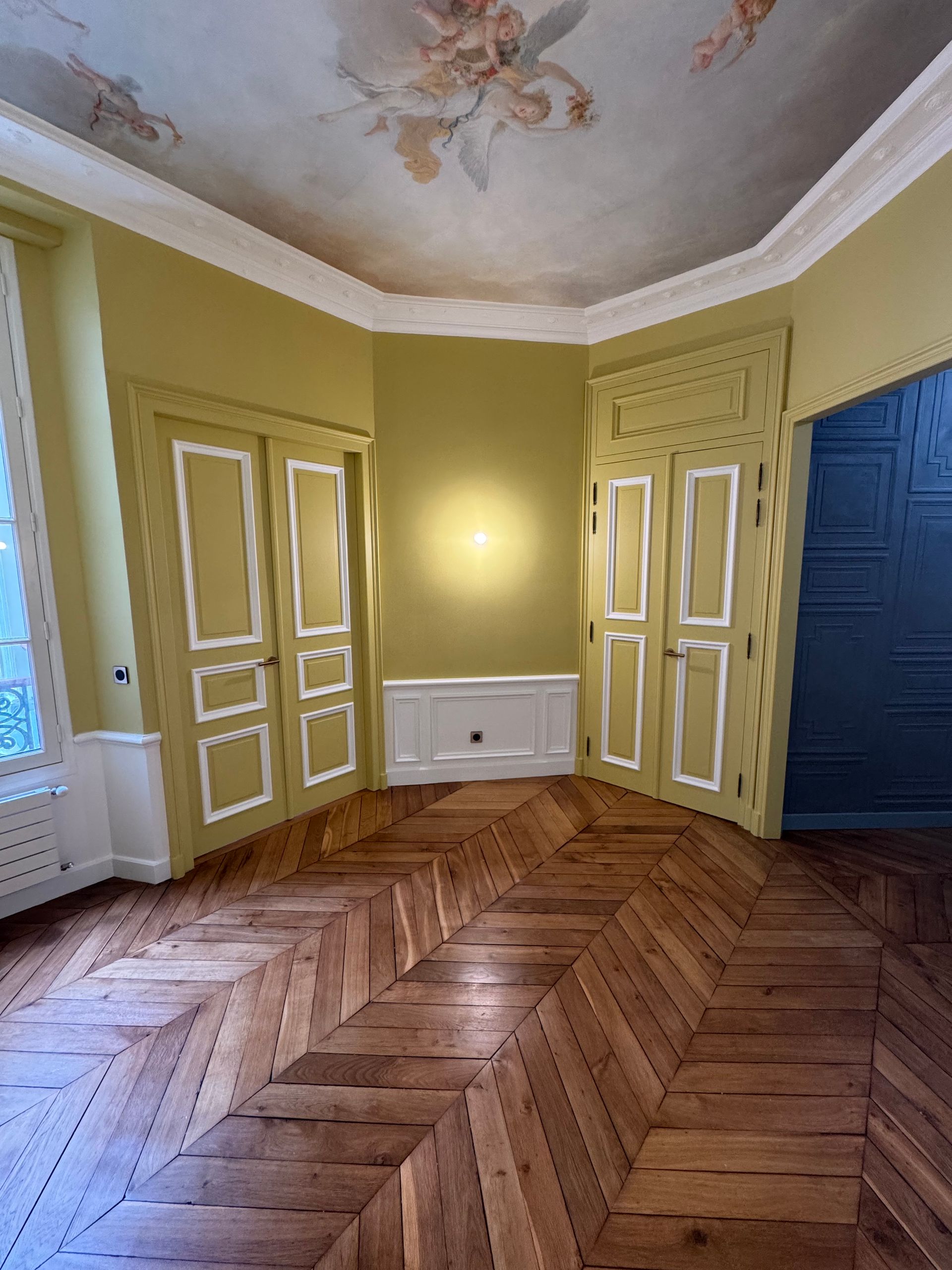 Pièce avec parquet, murs jaunes, plafond décoré et deux portes ornées.