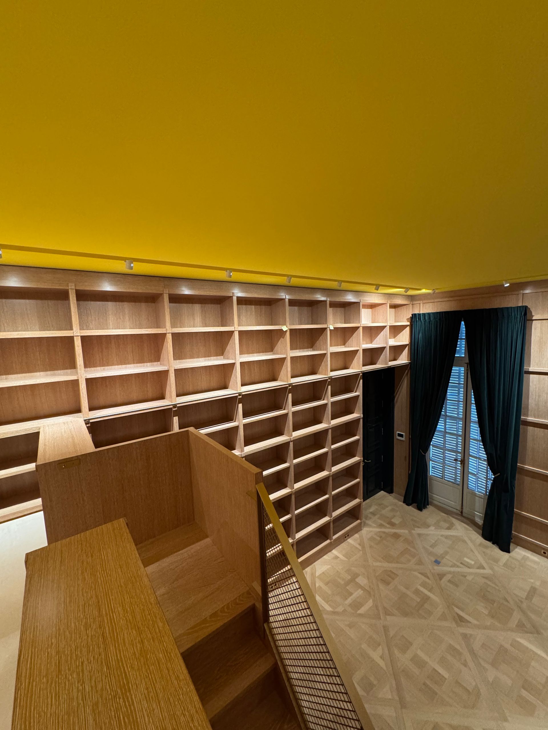 Intérieur d'une bibliothèque en bois avec des étagères vides, un escalier et une fenêtre aux rideaux sombres. Plafond jaune.