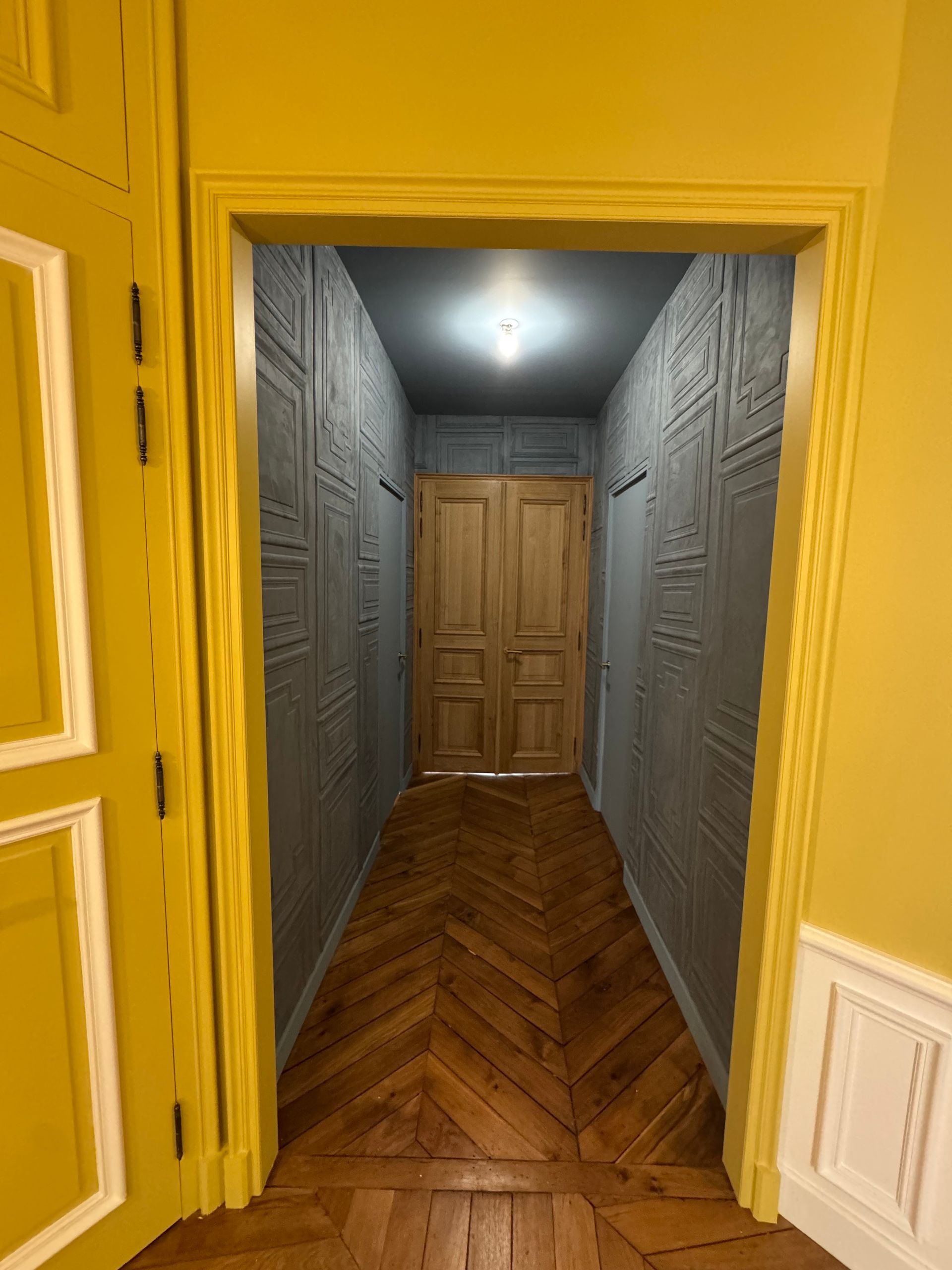 Un cadre de porte jaune donne sur un couloir étroit aux murs gris texturés, au sol en bois et éclairé au fond.